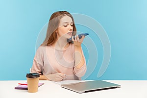 Digital speaker app. Young woman employee sitting at workplace and talking to mobile phone, using voice assistant photo