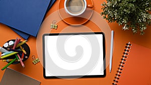 Digital tablet and stylus pen surrounded by office supplies on orange background.