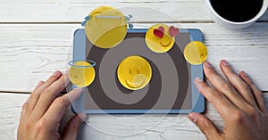 Digitally generated image of emojis flying over hands using digital tablet by coffee cup at table