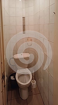dilapidated toilet in a small bathroom, walls covered with old tiles falling off