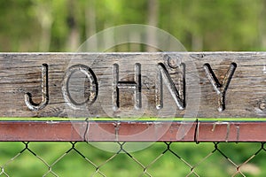 Dilapidated vintage retro sign with carved Johny on top mounted on metal pole of wire fence with green garden in background