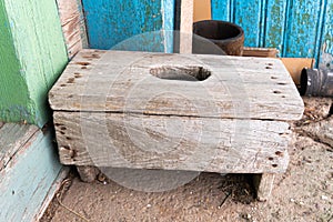 A dilapidated wooden stool with a hole in the seating area on the concrete floor near the village house