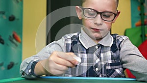 A diligent Caucasian boy in glasses of 5-7 years old writes in chalk on a blackboard, deciding homework in a home setting.