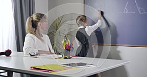 Diligent Caucasian schoolgirl turning to blackboard and writing with chalk. Adult woman sitting at the table and