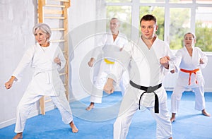 Diligent elderly women and men attendee of karate classes practicing kata standing in row with others in sports gym