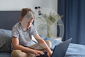 Diligent schoolboy using his laptop for doing homework, sitting on his bed in modern interior. Studying online during