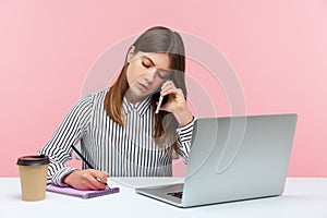 Diligent secretary in striped shirt receiving calls on phone and making notes, writing down information, sitting at workplace with