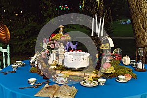 The dining table is decorated in the style of Alice in Wonderland. An open old book, a cake, tea bowls, candles in a