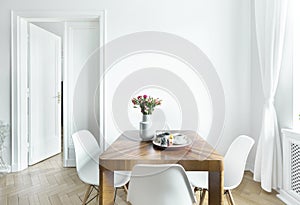 Dining table with fresh flowers and breakfast tray with coffee cup and fruits in real photo of white room interior with empty wall