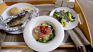 Dinner fish, salad, rice and boiled vegetables on wooden tray in hotel room.