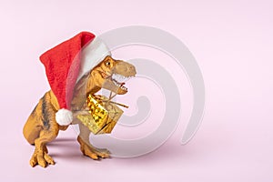 Dinosaur Rex in red Santa Claus hat holds golden gift box in its paws on pink background New Years Eve or Christmas Eve Art