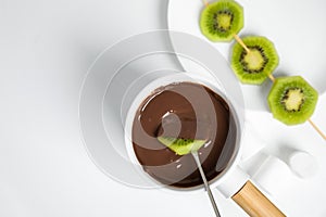 Dipping kiwi into fondue pot  chocolate on white table, flat lay. Space for text