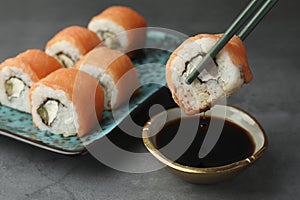 Dipping tasty sushi roll with salmon into soy sauce on grey table, closeup