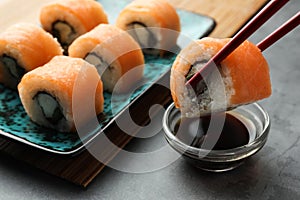 Dipping tasty sushi roll with salmon into soy sauce on grey table, closeup