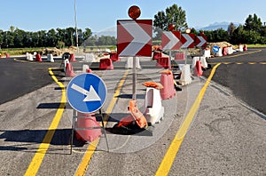 Directional road signs with warning lights and red and white plastic barriers in front of a roundabout under construction.