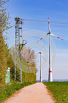 A dirt road at the edge of a field with wind turbines for power generation and a terminal pylon in Germany