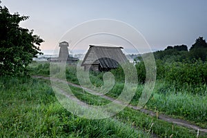 A dirt road through a field along an abandoned barn and an old mill. Early morning with fog in summer