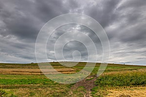 Dirt road leading to the top of a green hill at summertime, gathering storm clouds