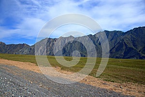 A dirt road running through a pasture with fresh grass at the foot of a high hill under a blue sky with white cumulus clouds