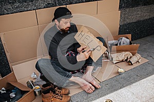 Dirty homeless man is sitting on cardboard and writing down the word help on a piece of paper. He has a lot of stuff on