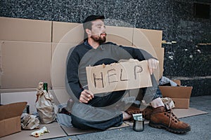 Dirty man is sitting on the ground and holding a help cardboard in hands. He is looking to the side. There are lots of