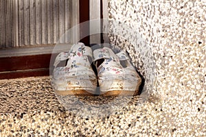 Dirty old woman shoes with flowers painted on white surface left in front of family house entrance doors at sunset