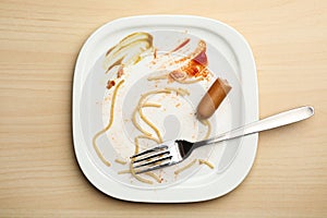 Dirty plate with food leftovers, sausage and fork on wooden background