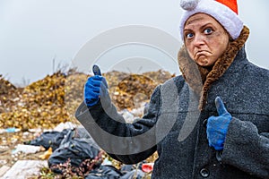 Dirty poor homeless woman in a New Year`s hat, unsure of herself, shows a thumb up class against the background of a dump