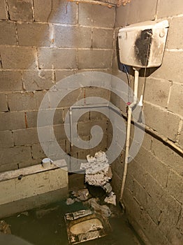 Dirty toilet room with pile of used toilet paper in the corner, uncoated concrete flooring and cinder brick walls.