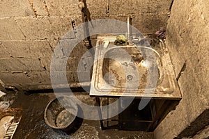 Dirty toilet room stainless steel sink with a bucket on the floor, uncoated concrete flooring and cinder brick walls.