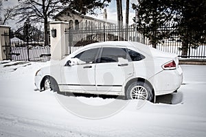 Dirty white car with scuffs parked in deep  snow on residential street outside tall fence with luxury mansion and estate in