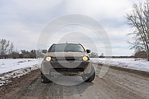 A dirty white SUV crossover stands with its headlights on before driving on a winter village.