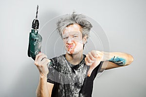 Dirty young builder guy in plaster is hold a green drill perforator on gray background at home during repairs