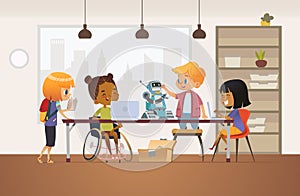 Disabled African American girl in wheelchair and other children standing around desk with laptops and robot and working