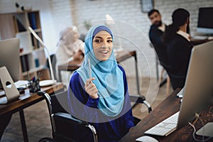 Disabled arab woman in wheelchair working in office. Woman is working on desktop computer.