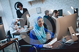 Disabled arab woman in wheelchair working in office. Woman is working on desktop computer and drinking coffee.