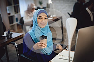 Disabled arab woman in wheelchair working in office. Woman is working on desktop computer and drinking coffee.