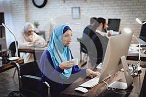 Disabled arab woman in wheelchair working in office. Woman is working on desktop computer and drinking coffee.