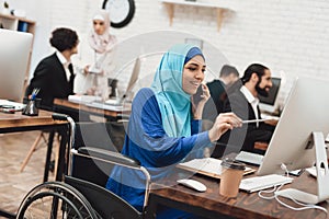 Disabled arab woman in wheelchair working in office. Woman is working on desktop computer and talking on phone.