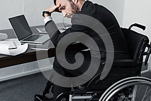 Disabled businessman sitting at workplace in wheelchair and holding hand on forehead