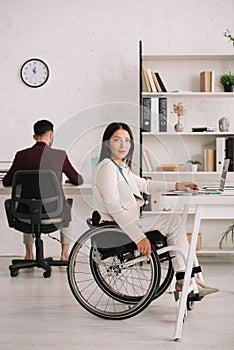 Disabled businesswoman looking at camera while sitting in wheelchair near colleague
