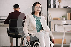 Disabled businesswoman looking at camera while sitting in wheelchair near colleague working on background