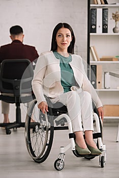 Disabled businesswoman smiling at camera while sitting in wheelchair near colleague working on background