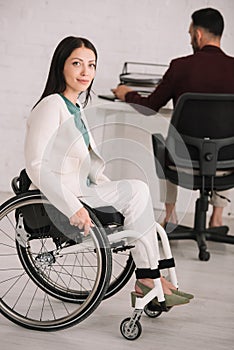 Disabled businesswoman smiling at camera while sitting in wheelchair near colleague working on background