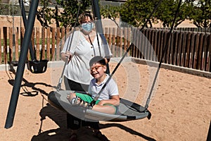 Disabled child in swing set with mother wearing a pandemic coronavirus protective mask