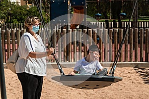 Disabled child in swing set with mother wearing a pandemic coronavirus protective mask