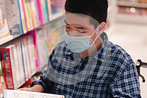 Disabled child on wheelchair wearing a mask reading a book from shelves in books store or school library
