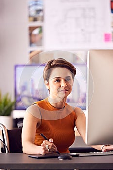 Disabled Female Architect In Wheelchair In Office Working On Desktop Computer Using Graphics Tablet