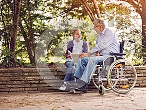 Disabled grandfather in wheelchair in park spending time together with his granddaughter reading book.