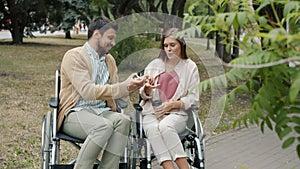 Disabled guy talking to handicapped girl outdoors taking her smartphone sitting in wheelchairs enjoying park